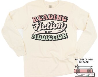 May include: A cream colored long-sleeved t-shirt with the text "Reading Fiction is my Addiction" in a retro font with pink, green, and black colors. The text is on the front of the shirt. The back of the shirt has a stack of books with stars and the text "Full size design on back".