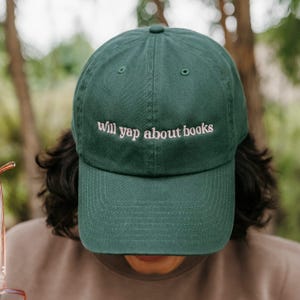 May include: A dark green baseball cap with the embroidered text "will yap about books" in a light pink color. The cap has a curved brim and a classic six-panel design. The hat is made of a soft material.
