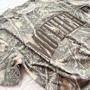 Op de afbeelding: Een t-shirt met korte mouwen met een camouflagepatroon en het woord "WILDERNESS" in een bruin, verhoogd lettertype over de borst. De shirt heeft een mix van groene, bruine en bruine kleuren, waardoor een boscamouflage-ontwerp ontstaat. De stof lijkt zacht en licht.