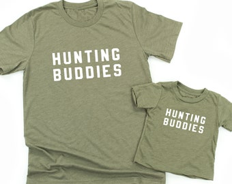 May include: Two olive green t-shirts with the text "Hunting Buddies" in white. The larger shirt is for an adult and the smaller shirt is for a child.