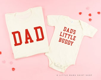 May include: Two white t-shirts with red text. The larger shirt says "DAD" and the smaller shirt says "DAD'S LITTLE BUDDY".