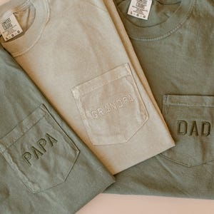 May include: Three Comfort Colors t-shirts in shades of green and tan. Each shirt has a pocket with embroidered text: "PAPA", "GRANDPA", and "DAD". The shirts are folded, showcasing the pocket details.