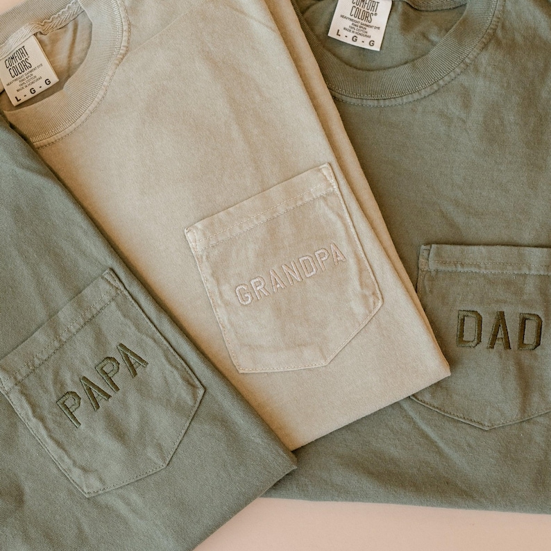 May include: Three Comfort Colors t-shirts in shades of green and tan. Each shirt has a pocket with embroidered text: "PAPA", "GRANDPA", and "DAD". The shirts are folded, showcasing the pocket details.
