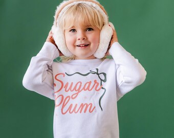 Può includere: Un bambino indossa una maglietta bianca a maniche lunghe con le parole "Sugar Plum" in corallo e un disegno a fiocco verde. Il bambino indossa anche paraorecchie color crema con finiture marroni, su sfondo verde.