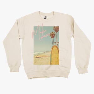 May include: A beige sweatshirt featuring a graphic print of a surfboard on a beach with palm trees. The text "Make Waves" is written in pink on the graphic.
