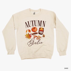 May include: Sand-colored sweatshirt with the words "AUTUMN Girlie" in brown script. The graphic includes a pumpkin, fall leaves, a cinnamon stick, and a candle. The sweatshirt is a crew neck style.