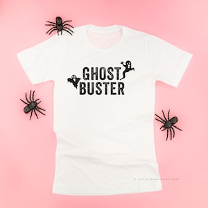 May include: White crew neck t-shirt featuring the text "GHOST BUSTER" in black, with three ghost graphics. The shirt is surrounded by black plastic spiders on a pink backdrop. A Halloween-themed garment.