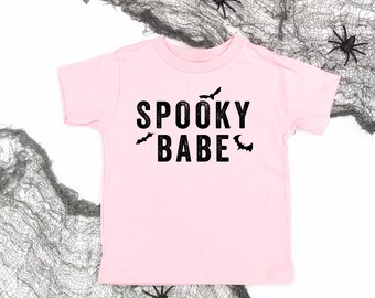 Könnte beinhalten: Ein hellrosa T-Shirt mit den schwarzen Worten "SPOOKY BABE" und Fledermausgrafiken. Das Shirt ist mit schwarzen Spinnen und einem grauen spinnennetzartigen Stoff dekoriert, perfekt für Halloween.