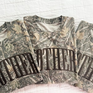 Op de afbeelding: Drie opgevouwen camouflage T-shirts met het woord "MOTHER" gedrukt in donkerbruine blokletters. De shirts hebben een ronde hals en zijn gemaakt van een camouflagepatroon in tinten groen, bruin en beige.