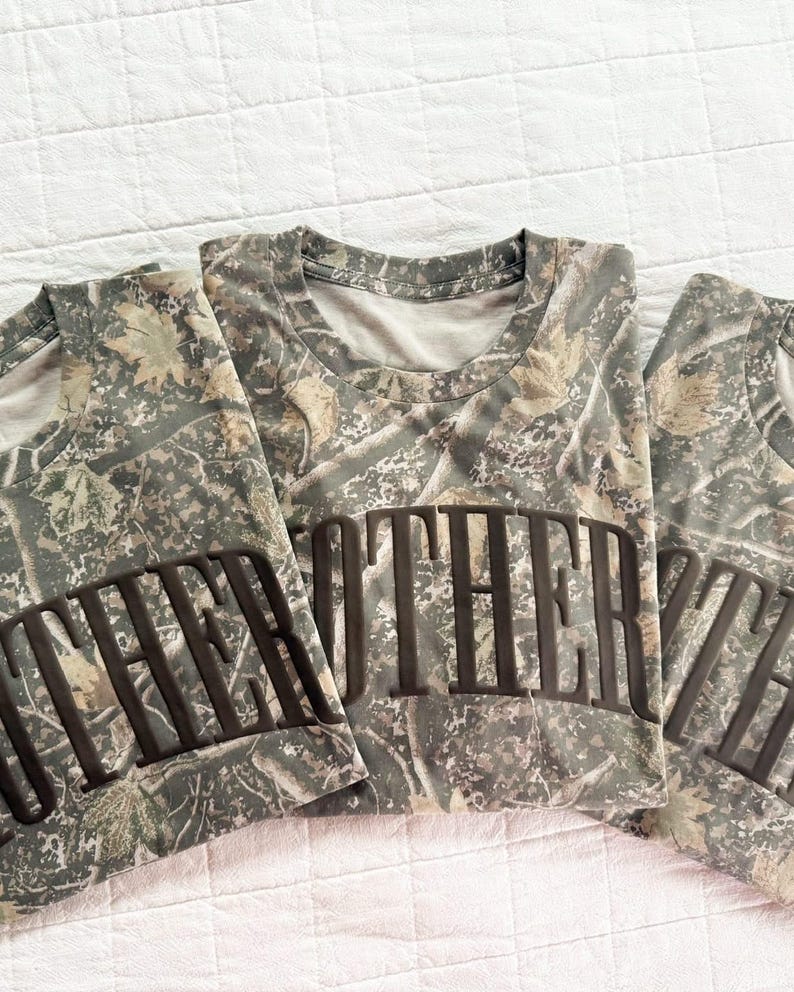 Op de afbeelding: Drie opgevouwen camouflage T-shirts met het woord "MOTHER" gedrukt in donkerbruine blokletters. De shirts hebben een ronde hals en zijn gemaakt van een camouflagepatroon in tinten groen, bruin en beige.