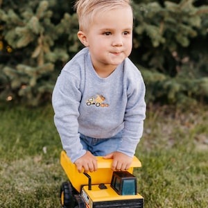 May include: A young child wearing a gray sweatshirt with a small embroidered construction truck design. The child is standing on a large yellow and black toy dump truck on a grassy lawn.
