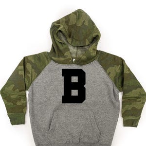 May include: Gray hoodie with a black letter "B" on the front. The sleeves and hood are camouflage print in shades of green, brown, and tan. The hoodie has a front pocket and a drawstring hood.