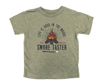 Op de afbeelding: Een groen T-shirt met een kampvuur-afbeelding en de tekst "Life is good in the woods. The official 'S'more Taster."