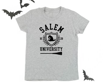 Könnte beinhalten: Hellgraues T-Shirt mit schwarzem Aufdruck. Das Design zeigt die Worte "Salem University" in einem Schild, einen Hexenhut, die Zahlen "16" und "29" und einen Besen. Dekorative Fledermaus-Ausschnitte befinden sich im Hintergrund.