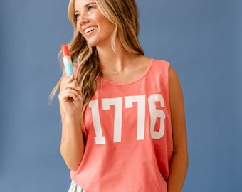 May include: A coral-coloured tank top with the number "1776" printed in white. The person is holding a red, white, and blue ice lolly. The background is a solid blue.