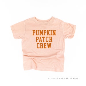 Puede incluir: Camiseta de color melocotón claro con la frase "PUMPKIN PATCH CREW" en naranja intenso. Esta camiseta de manga corta y cuello redondo es ideal para niños. El texto está centrado en la parte delantera de la prenda.