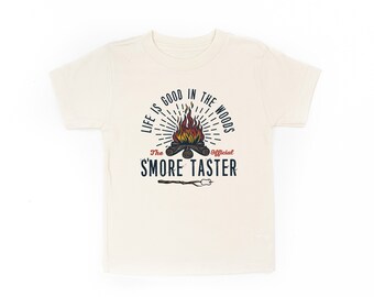 Op de afbeelding: Een beige t-shirt met een kampvuur-afbeelding en de tekst "Life is good in the woods. The Official 'S'more Taster."