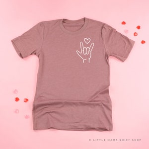 May include: A mauve colored t-shirt with a white outline of the American Sign Language symbol for "I love you" with a small heart above the hand.