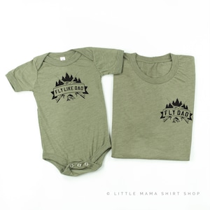 Op de afbeelding: Twee groene shirts met een zwarte afbeelding van gekruiste hengels, een vis en bomen. Het grotere shirt zegt "Fly Dad" en het kleinere shirt zegt "Fly Like Dad".