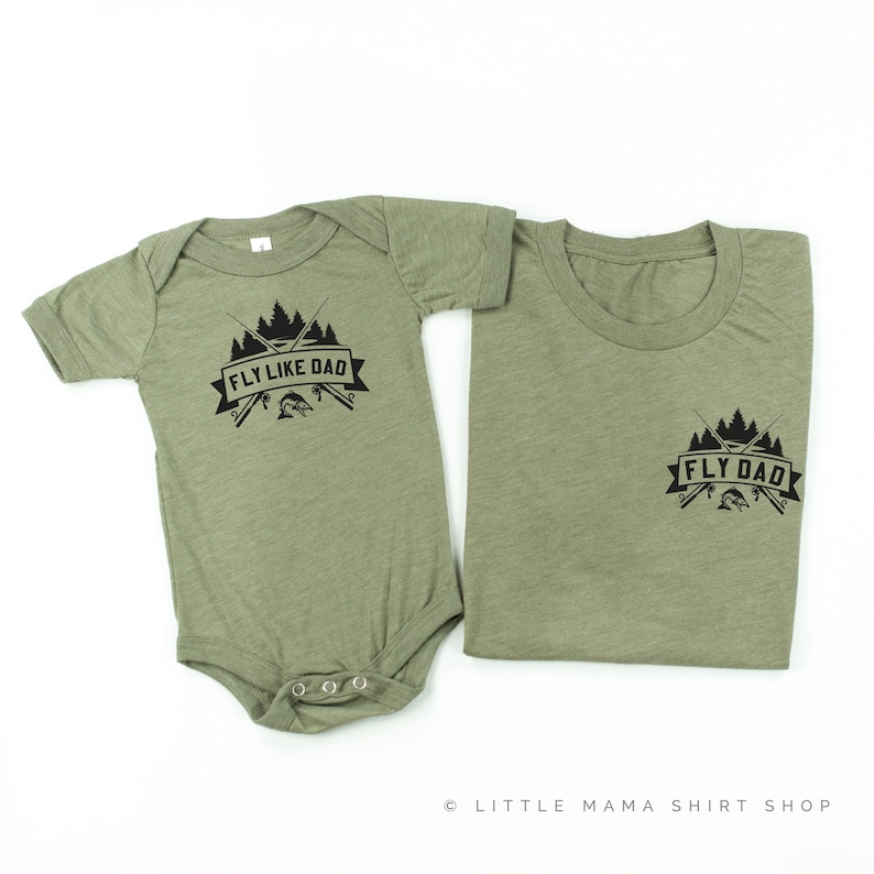 Op de afbeelding: Twee groene shirts met een zwarte afbeelding van gekruiste hengels, een vis en bomen. Het grotere shirt zegt "Fly Dad" en het kleinere shirt zegt "Fly Like Dad".
