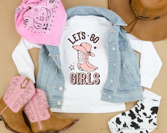 Puede incluir: Camiseta blanca de manga larga con el texto "Let's Go Girls" y un gráfico de botas de vaquero rosas. La camiseta se combina con una chaqueta vaquera, un pañuelo rosa, un sombrero de vaquero marrón y calcetines de vaca en blanco y negro.