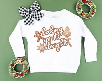 Può includere: Felpa bianca a maniche lunghe con la frase "baking spirits bright" in marrone e disegni di biscotti di pan di zenzero. Un fiocco a scacchi bianco e nero e due ghirlande decorative con ornamenti rossi, verdi e dorati sono presenti nell'immagine.