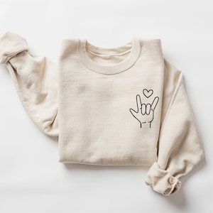 May include: A beige sweatshirt with a black outline of the American Sign Language symbol for "I love you" with a small heart above it.
