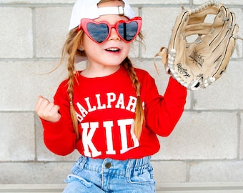 May include: A young girl wearing a red long-sleeved shirt with the text "Ballpark Kid" in white letters, a white baseball cap, heart-shaped sunglasses, and denim shorts. She is holding a brown baseball glove.