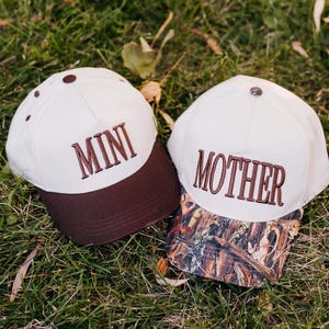 Puede incluir: Dos gorras de béisbol. Una con la palabra "MINI" bordada en marrón y visera marrón, la otra con "MOTHER" y visera de camuflaje. Ambas gorras son blancas y están sobre hierba.