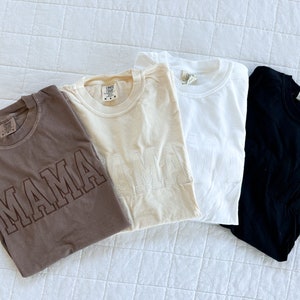 May include: Three t-shirts with the word "MAMA" embroidered on them. The shirts are brown, beige, and white.
