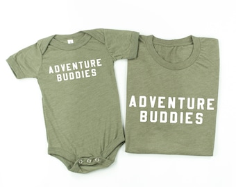 May include: Two green shirts with the text "Adventure Buddies" in white. The smaller shirt is a baby onesie and the larger shirt is a t-shirt.