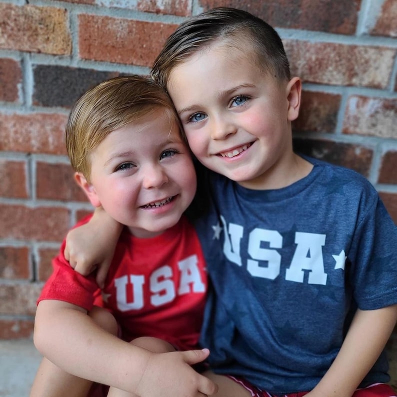 May include: Two young boys wearing red and blue t-shirts with the word "USA" printed on them. They are smiling and standing in front of a brick wall.
