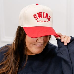May include: A white baseball cap with a red brim. The front of the cap has red embroidered text that reads "Swing Batter Batter".