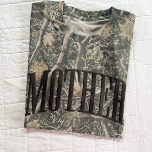 Op de afbeelding: T-shirt met camouflagepatroon met het woord "MOTHER" in grote, donkere letters over de voorkant. Het shirt heeft een ronde hals en een mix van groene, bruine en bruine kleuren, waardoor een bos-camouflage-ontwerp ontstaat.
