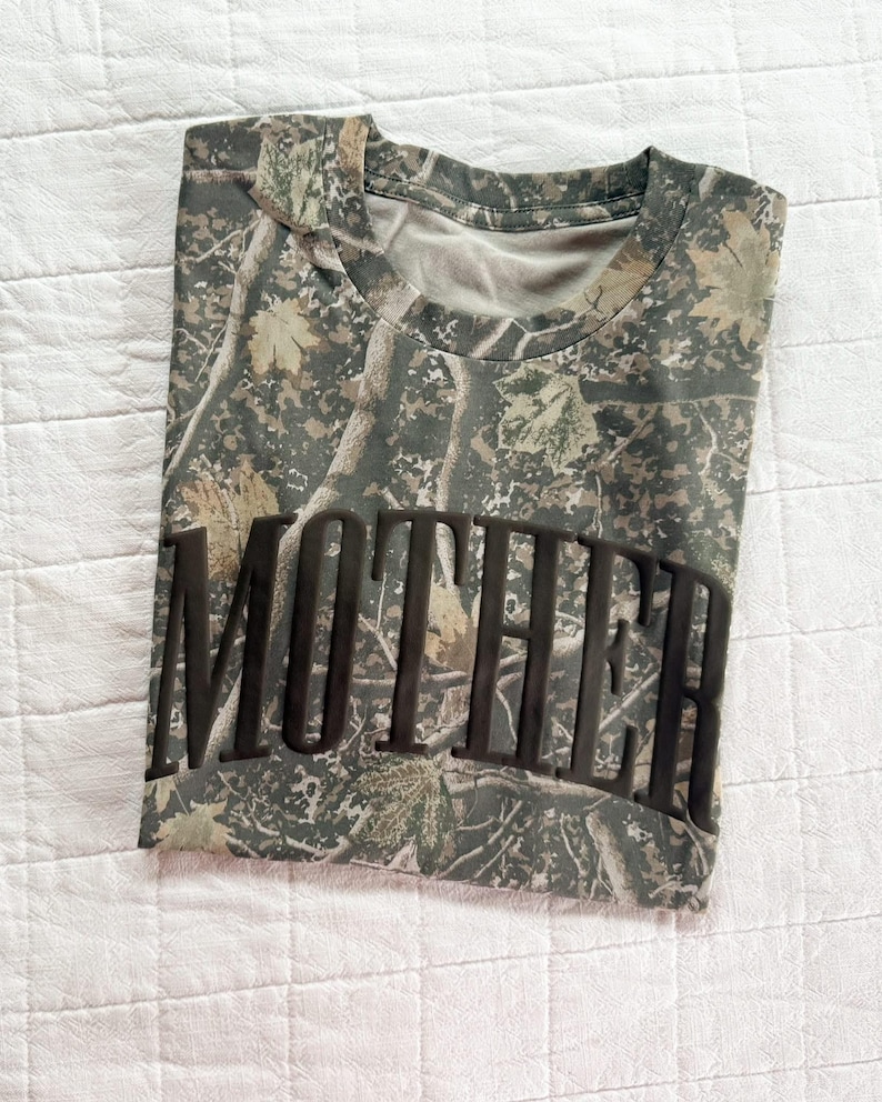 Op de afbeelding: T-shirt met camouflagepatroon met het woord "MOTHER" in grote, donkere letters over de voorkant. Het shirt heeft een ronde hals en een mix van groene, bruine en bruine kleuren, waardoor een bos-camouflage-ontwerp ontstaat.