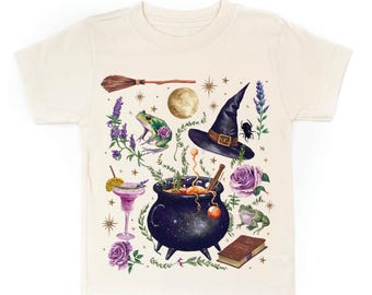 May include: Cream-colored t-shirt featuring a whimsical witch-themed design. The graphic includes a black cauldron, witch's hat, broom, moon, frog, flowers, and a cocktail. The design is in shades of purple, green, brown, and black.
