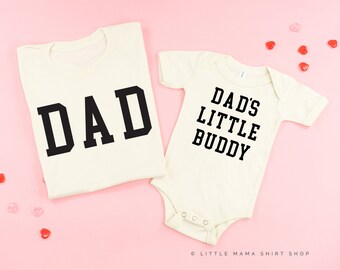 Könnte beinhalten: Zwei cremefarbene Shirts mit schwarzem Text. Das größere Shirt sagt "DAD" in großen Blockbuchstaben. Das kleinere Shirt sagt "DAD'S LITTLE BUDDY".