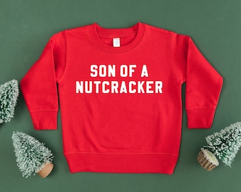 May include: A red sweatshirt with the text "SON OF A NUTCRACKER" in white block letters. The sweatshirt is surrounded by small, decorative Christmas trees on a green background.