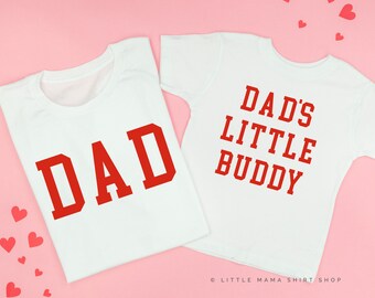 May include: Two white t-shirts with red text. The larger shirt says "DAD" in large, bold letters. The smaller shirt says "DAD'S LITTLE BUDDY".
