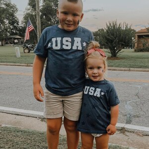 May include: Two children wearing matching navy blue t-shirts with white stars and the word "USA" printed on the front. The boy is wearing khaki shorts and the girl is wearing a white diaper cover. They are standing on a sidewalk in front of a grassy area.