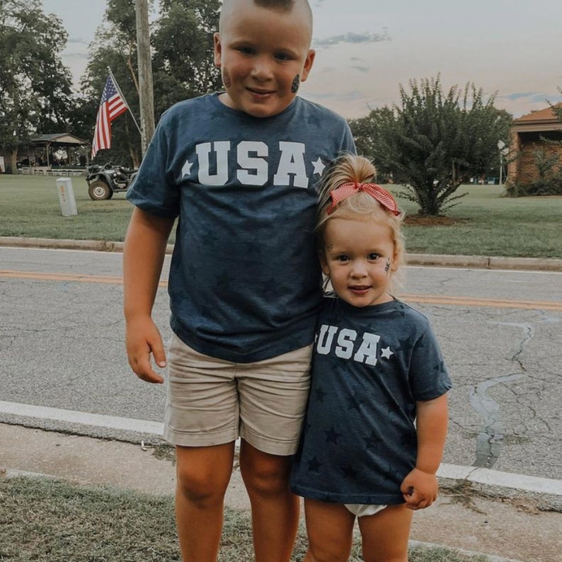May include: Two children wearing matching navy blue t-shirts with white stars and the word "USA" printed on the front. The boy is wearing khaki shorts and the girl is wearing a white diaper cover. They are standing on a sidewalk in front of a grassy area.