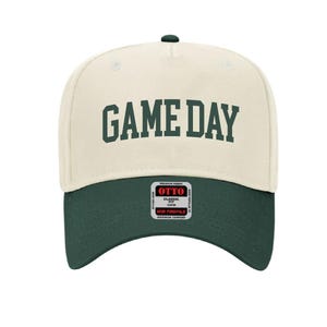 May include: A cream colored baseball cap with a green brim. The front of the cap has the text "GAME DAY" in green.