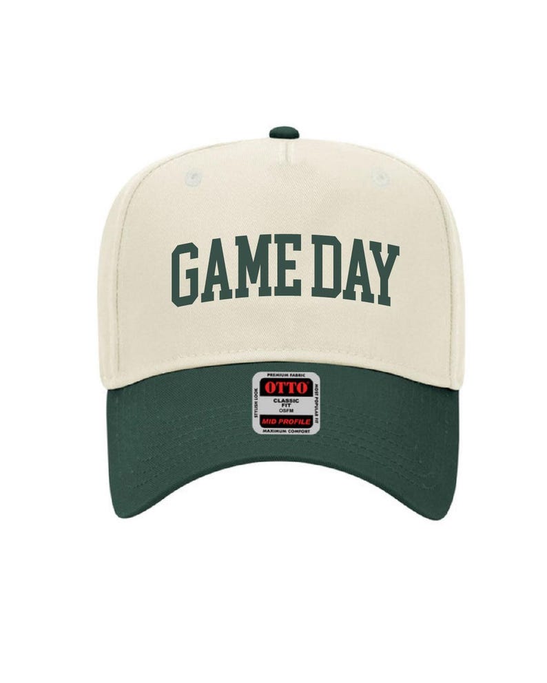 May include: A cream colored baseball cap with a green brim. The front of the cap has the text "GAME DAY" in green.
