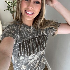 Op de afbeelding: Een camouflage-patroon T-shirt met het woord "MOTHER" in bruine blokletters over de borst. Het shirt heeft een ronde hals en korte mouwen. De achtergrond is een mix van groen, bruin en beige.