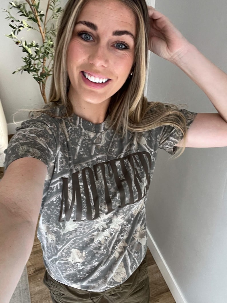 Op de afbeelding: Een camouflage-patroon T-shirt met het woord "MOTHER" in bruine blokletters over de borst. Het shirt heeft een ronde hals en korte mouwen. De achtergrond is een mix van groen, bruin en beige.