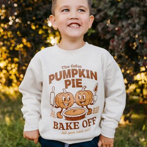 May include: A child wearing a beige sweatshirt with a brown graphic that reads "The Great Pumpkin Pie Bake Off." The graphic features two cartoon pumpkins and a pie. The child is also wearing navy blue pants.
