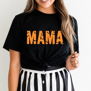 May include: Black t-shirt featuring the word "MAMA" in orange block letters, with black bat silhouettes within. The shirt has a Halloween theme and is knotted at the waist.
