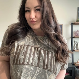 Op de afbeelding: Een camouflage-patroon T-shirt met het woord "WHITTON" in bruine blokletters. Het shirt heeft korte mouwen en een ronde hals. Het kleurenschema is een mix van groen, bruin en beige.