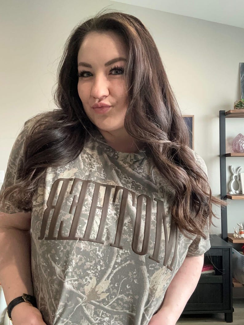 Op de afbeelding: Een camouflage-patroon T-shirt met het woord "WHITTON" in bruine blokletters. Het shirt heeft korte mouwen en een ronde hals. Het kleurenschema is een mix van groen, bruin en beige.