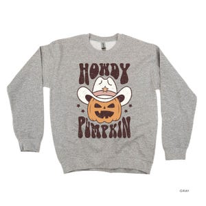Könnte beinhalten: Ein graues Sweatshirt mit einer Grafik einer Kürbis-Figur mit Cowboyhut. Der Text lautet "HOWDY PUMPKIN" in braunen Buchstaben. Der Kürbis ist orange mit einem geschnitzten Gesicht, und der Hut ist weiß.